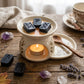 A styled tabletop scene with a white ceramic candle warmer holding a lit tealight candle and three black wax melts with runic symbols. The setup includes dried lavender, crystals, autumn leaves, a teacup and saucer, and a cloth on a rustic wooden surface.